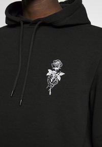 Sweat à capuche noir avec cordons, présentant un motif brodé blanc d'une main squelettique tenant une rose avec des feuilles au niveau de la poitrine.