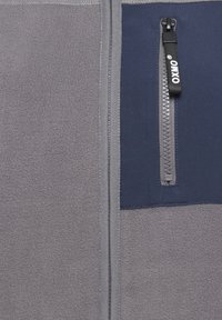 Giacca in pile grigia con un pannello blu navy, dotata di chiusura con zip, cuciture a contrasto e una tasca con zip sul lato sinistro del petto.