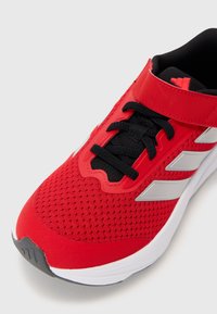 Red athletic shoe featuring a breathable mesh upper, black accents, a Velcro strap, and silver side stripes. Rubber sole with a textured finish.