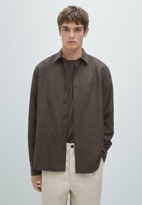 Brown button-up shirt with a textured fabric, long sleeves, and a relaxed fit, paired with a dark t-shirt and light-colored trousers.