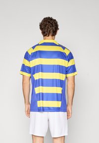 Mand, der vender væk, iført en blå og gul horisontalt stribet sportsjersey og hvide shorts mod en ensfarvet baggrund.