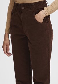 Brown corduroy pants with an elasticated waistband, a button closure, and side pockets. The fabric has a soft texture and visible ribbing pattern.