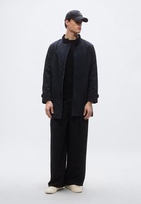 Μαύρο, ελαφρύ παλτό με κούμπωμα με κουμπιά και ψηλό γιακά· συνδυασμένο με μαύρο πουλόβερ και άνετο μαύρο παντελόνι. Λευκά παπούτσια.