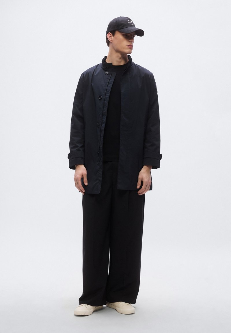 Black, lightweight coat with button closure and high collar; paired with black sweater and relaxed-fit black trousers. White footwear.