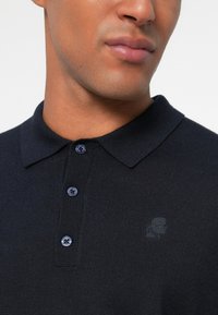 Námornícky modré polo tričko s textúrovaným pletením, tromi čiernymi gombíkmi a jemným logom v látke. Detailný pohľad na golier a horné gombíky.