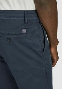 Marineblaue Cargo-Shorts mit glatter Textur, ausgestattet mit einer Gesäßtasche und einem Logo-Patch. Der Bund hat verstellbare Elemente.