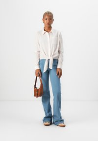 Lys rosa knyte skjorte i livet, kombinert med blå svingjeans. Modellen holder en liten brun håndveske. Glatte teksturer i stoffet.