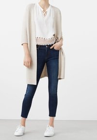Béžový dlouhý cardigan, bílá blůza s vázankou, tmavě modré skinny džíny a bílé tenisky. Cardigan má strukturovaný pletený vzor.