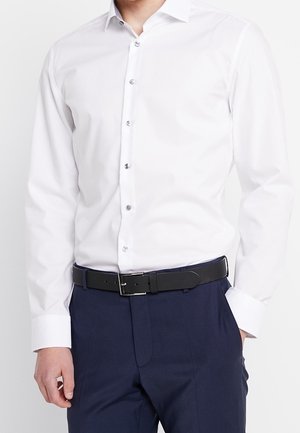 Mann trägt ein weißes langärmliges Hemd, das in marineblaue Hose mit schwarzem Gürtel gesteckt ist. Eine Hand in der Tasche.