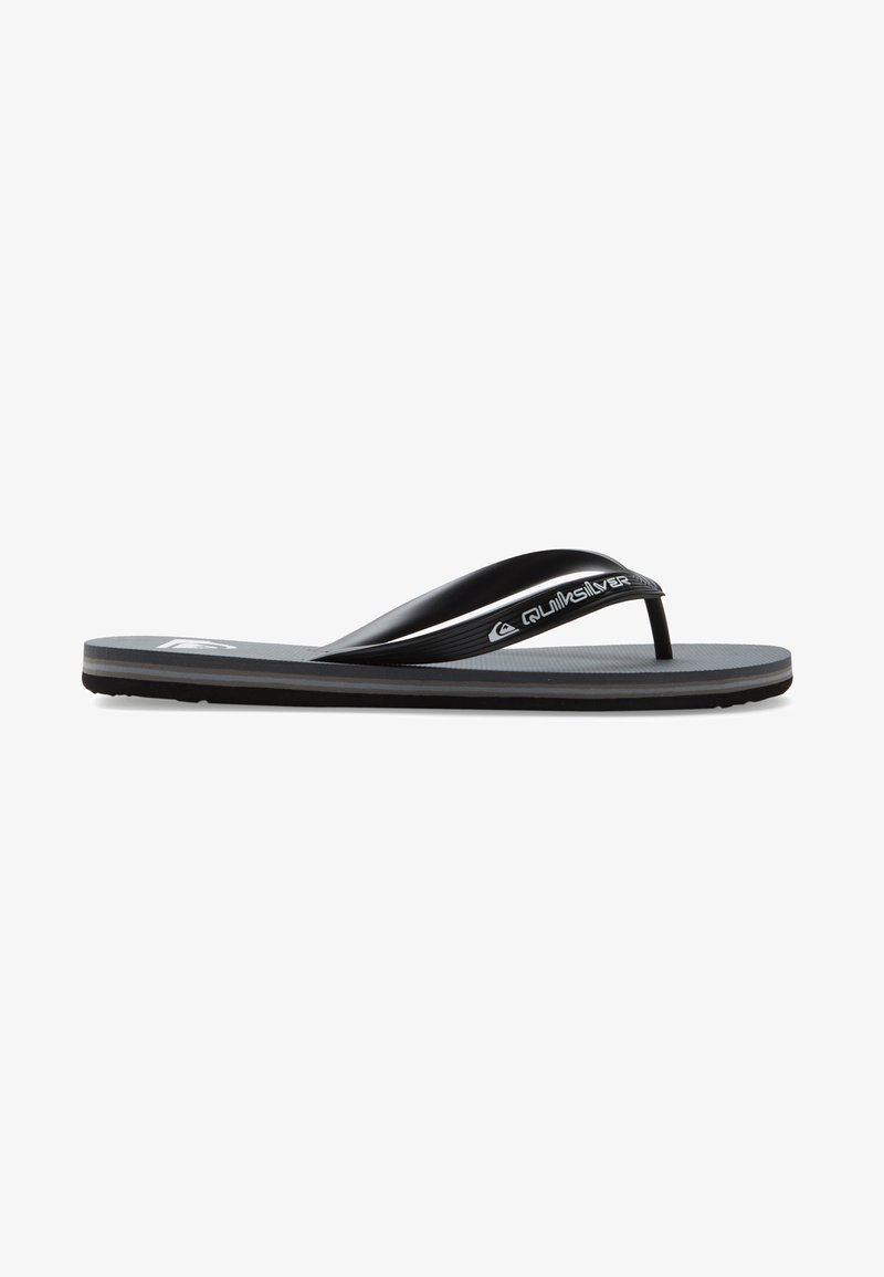 Svart flip-flop i gummi, med en texturerad sula, smala remmar och en logodetalj på remmen. Tunn yttersula med räfflor.