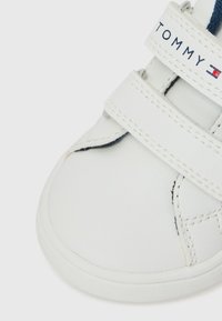 Hvit skinnsneaker med borrelåser, med blå detaljer og merkevarenavnet "TOMMY" trykket på stroppen. Glatt tekstur.