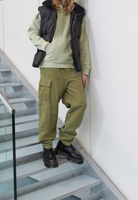 Gilet nero in piuma sopra un felpa verde, abbinato a pantaloni cargo larghi color oliva e stivali platform neri, in piedi su una scala con corrimano in vetro.