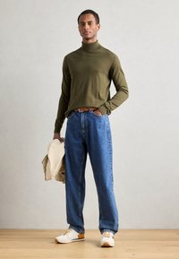 Olivově zelený rolák, modré volné džíny, béžová bunda v ruce, bílé tenisky s oranžovými akcenty, stojící na dřevěné podlaze.