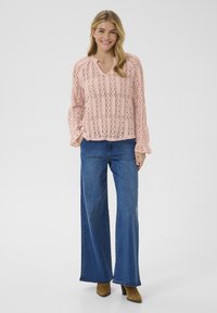 Svetlo roza čipkast bluza z V-izrezom in zvončastimi rokavi, v kombinaciji s širokimi jeanskimi hlačami v modri barvi in rjavimi gležnjarji.