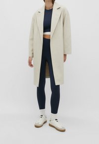 Osoba ubrana w beżowy płaszcz do kolan, granatowy krótki top i legginsy, białe trampki i skarpetki na jednolitym tle.