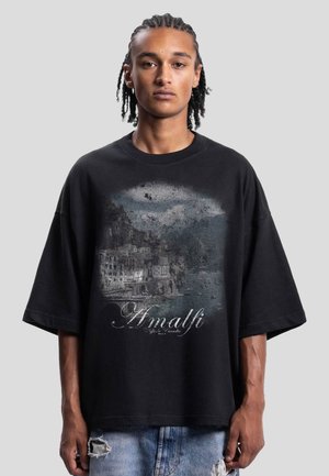 Young man with braided hair wearing a black oversized T-shirt with a faded Amalfi Coast landscape print and light ripped jeans.