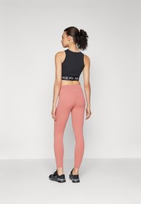 Svart avkortet tanktop med hvit logo, paret med tettsittende rosa leggings og svarte joggesko. Modell sett fra ryggen.