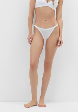 Femme portant un soutien-gorge blanc et une culotte blanche transparente, debout pieds nus devant un fond clair uni.