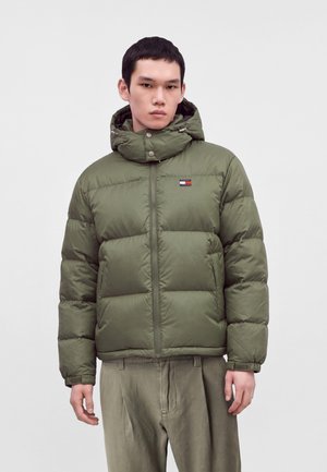 Jeune homme portant une doudoune verte avec capuche et fermeture éclair, associée à un pantalon vert ample, debout devant un fond blanc uni.