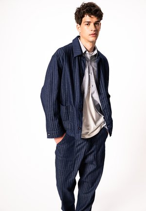 Mladi moški, oblečen v temno moder jopič z nežnimi črtami in ujemajoče hlače, preko sive srajce in bele majice, stoji z rokami v žepih.