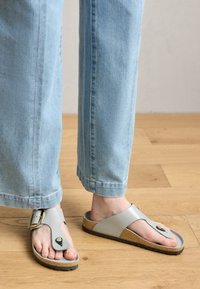Gray leather sandals with a toe post and a wide strap featuring a gold buckle. Cork footbed and black rubber sole. Worn with light blue jeans.