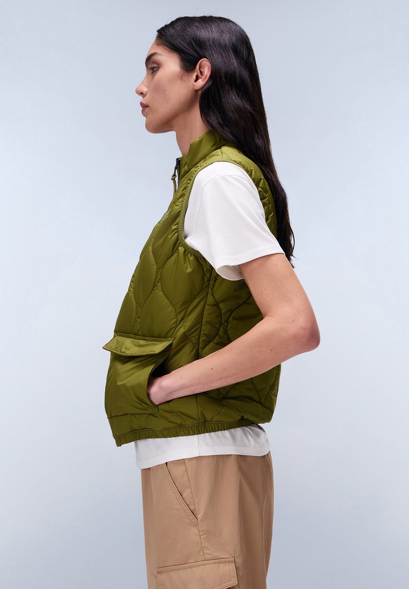 Zielona pikowana kamizelka bez rękawów z wysokim kołnierzem i kieszonką z przodu, zestawiona z białą koszulką i beżowymi spodniami, zaprezentowana z boku.