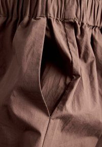 Καφέ ύφασμα με ελαστική μέση που μαζεύει και ανοιχτή πλαϊνή τσέπη που δείχνει την εσωτερική επένδυση του υφάσματος.