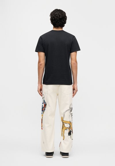 Μαύρο βαμβακερό μπλουζάκι συνδυασμένο με εκρού παντελόνι φαρδιάς γραμμής που διαθέτει πολύχρωμα καλλιτεχνικά σχέδια με φιγούρες στα πλάγια. Μαύρα παπούτσια ολοκληρώνουν την εμφάνιση.