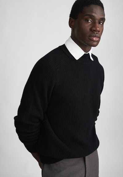 Pull noir côtelé avec un col rond, superposé à une chemise blanche à col. La texture semble douce et épaisse, offrant un look confortable.