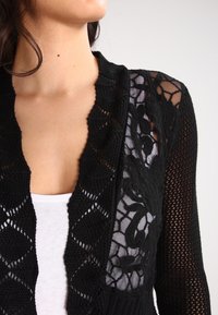 Cárdigan negro de punto abierto con un detalle de panel de encaje floral, patrones de diamante intrincados y un acabado texturizado.