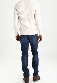 Pull crème en tricot avec motif torsadé texturé, assorti à un jean bleu foncé. Chaussures marron visibles, avec une tenue décontractée dans l'ensemble.