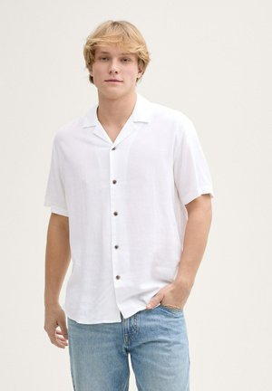 Chemise blanche à manches courtes avec col camp, cinq boutons marron, tissu léger, associée à un jean bleu clair.