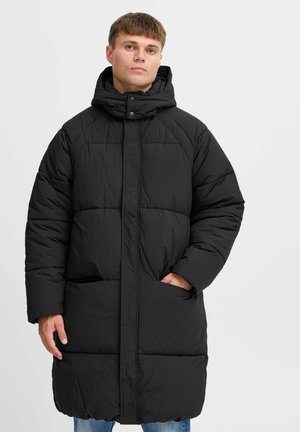 Svart, polstret lang kåpe med høy krage og trykknappslukking. Teksturert stoff og synlige quiltede seksjoner. Modellen viser frem lommer foran.