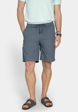 Hombre con camisa de manga corta azul claro, pantalones cortos grises con cordón y sandalias grises abiertas, de pie con las manos en los bolsillos.