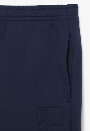 Pantaloni della tuta blu navy con vita elastica, tasche laterali e toppa con logo Lacoste Paris in rilievo vicino alla tasca.