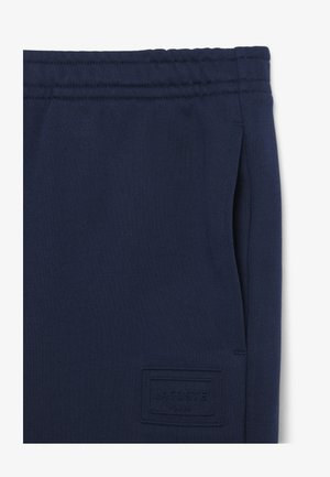 Pantaloni della tuta blu navy con vita elastica, tasche laterali e toppa con logo Lacoste Paris in rilievo vicino alla tasca.