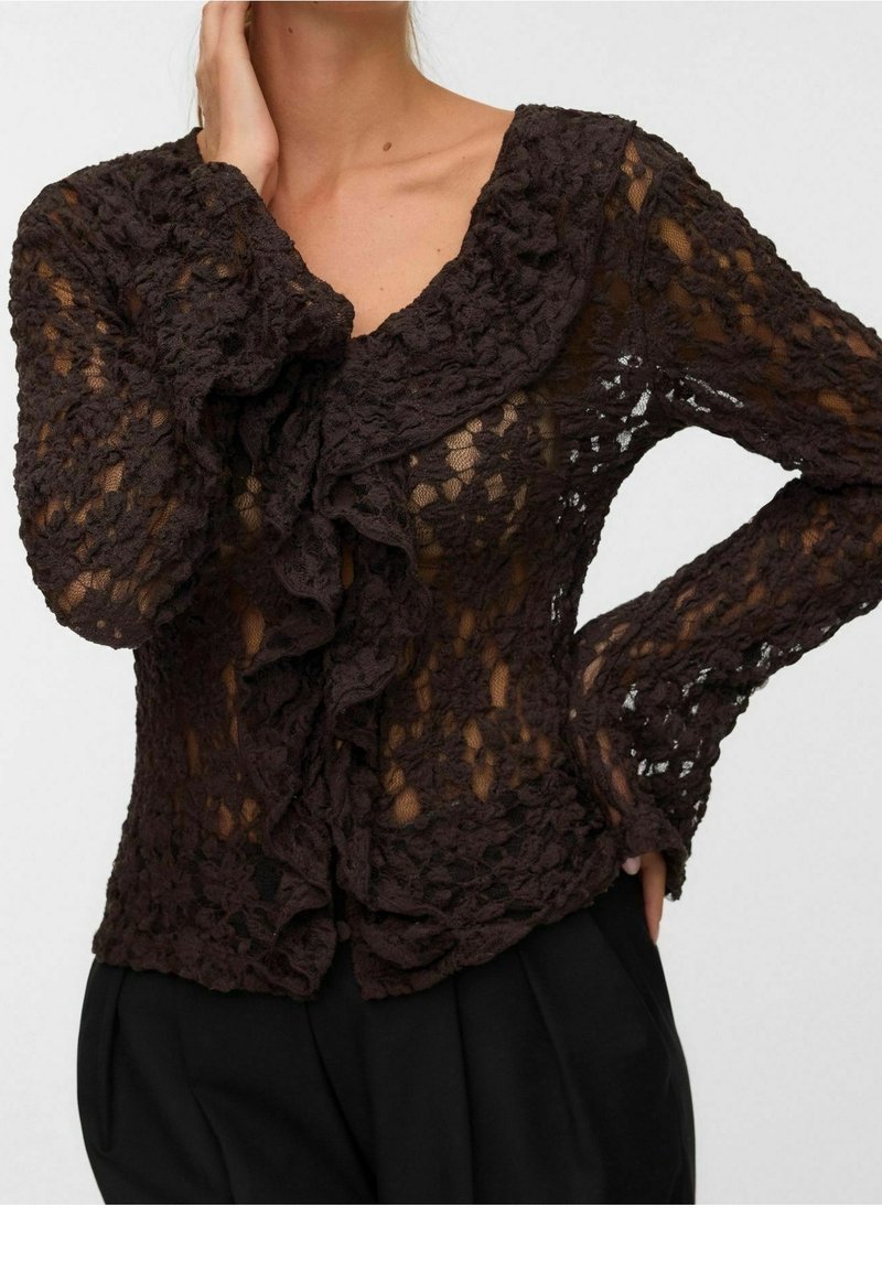 Femme portant un chemisier en dentelle fine marron foncé avec un devant à volants et de longues manches, associé à un pantalon noir sur un fond uni.