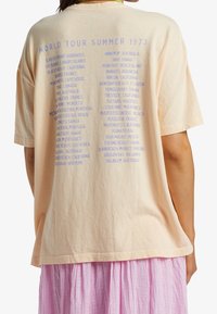 Perzikkleurig oversized t-shirt met blauwe tekst die locaties opsomt onder "WORLD TOUR SUMMER 1973." Zachte stof met een relaxte pasvorm.