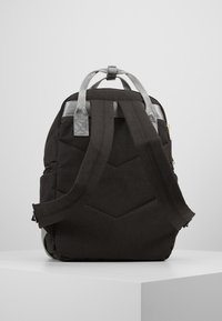 Sac à dos en tissu noir avec des bretelles rembourrées, une poignée en haut avec des accents argentés et un motif en chevrons matelassé à l'arrière. Il dispose d'une poche latérale.