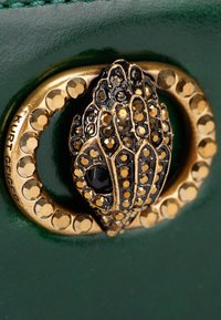 Versierde gouden en zwarte juwelen slangkopsluiting bevestigd aan donkergroen gestructureerd materiaal, ring versierd met gouden studs en letters.