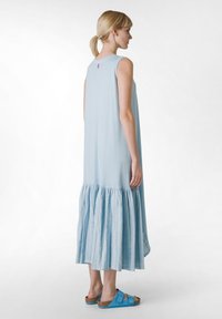 Rochie fără mâneci, de culoare albastru deschis, cu un tiv volănaș, realizată dintr-un material moale. Modelul poartă sandale albastre cu talpă plată. Vedere din spate.