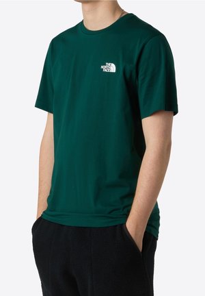 Mężczyzna w ciemnozielonej koszulce The North Face z krótkim rękawem i czarnych spodniach, ręce w kieszeniach, na białym tle.