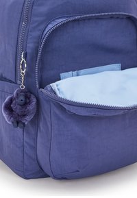 Lila ryggsäck med ett texturerat, hållbart material. Framfickan har en ljusblå foder och en fluffig lila nyckelringsanhängare.