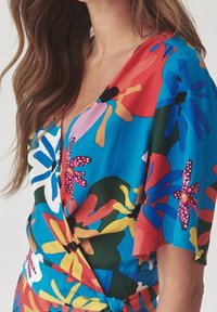 Värikäs kukkakuvioinen kietaisumekko, jossa suuret, näyttävät kukat punaisina, sinisinä, vaaleanpunaisina ja vihreinä turkoosilla taustalla, lyhyillä hihoilla.