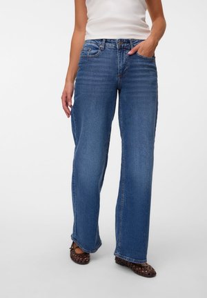 Femme debout portant un haut blanc sans manches, un jean bleu taille haute à jambes larges, et des chaussures noires ouvertes à l'avant, sur un fond clair uni.