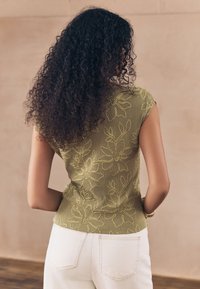 Top verde oliva con un design a maglia testurizzata, con ricami floreali in giallo. Maniche corte, stile aderente, abbinato a pantaloni bianchi.