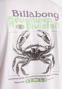 Λευκό βαμβακερό μπλουζάκι με μεγάλο μαύρο σχέδιο καβουριού. Το κείμενο σε μοβ και πράσινο γράφει "Billabong Adventure Division" και "Quality surf goods".