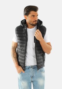 Gilet nero imbottito con cappuccio, caratterizzato da cuciture orizzontali, tasche con zip e chiusura frontale a zip, indossato sopra una camicia bianca.
