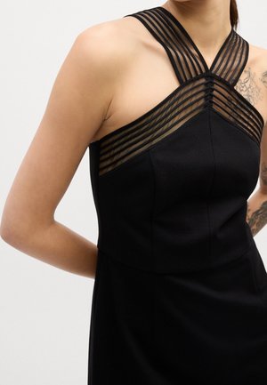 Mujer con un vestido negro sin mangas con tirantes transparentes a rayas que se cruzan en el pecho y tatuajes florales visibles en los brazos superiores.