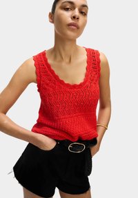 Rotes gehäkeltes Tanktop mit gewelltem Ausschnitt und Zickzackmuster, kombiniert mit schwarzen Shorts, die einen geflochtenen Gürtel und eine goldene Schnalle haben.